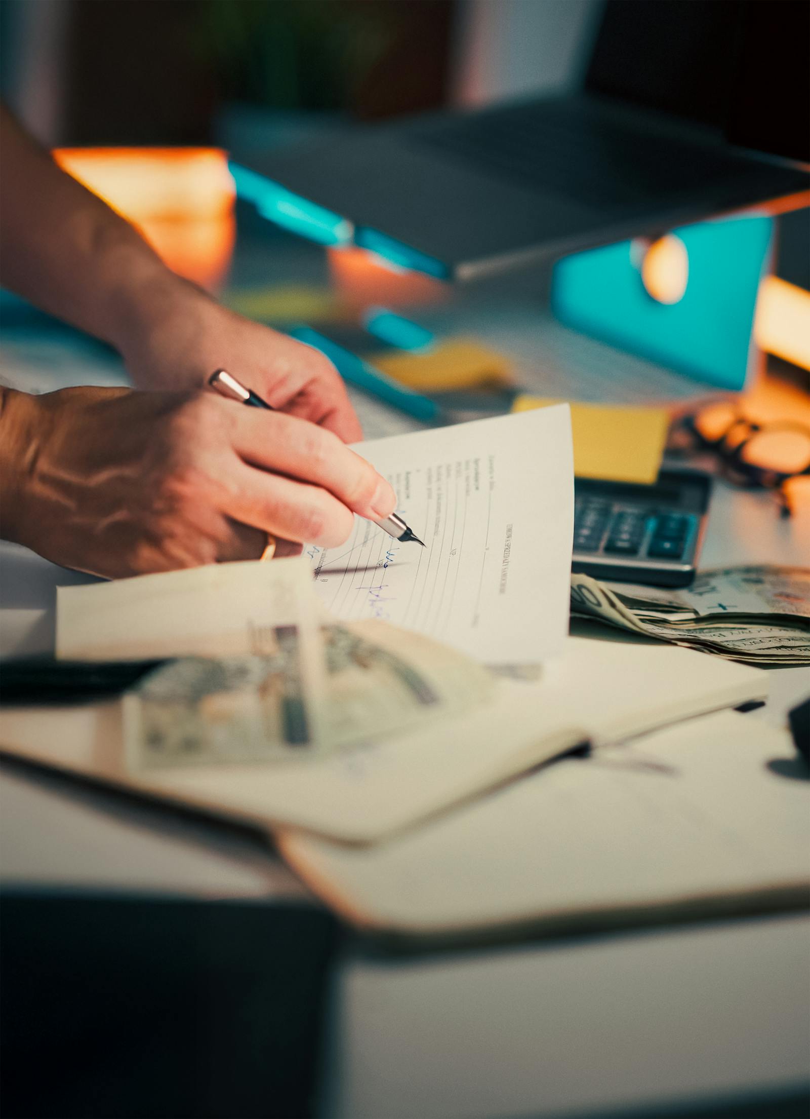 Hand signing a financial document with cash and a calculator on a moody desk